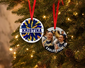 May include: Two round white ceramic ornaments with red ribbon hangers. One ornament features a blue and gold glitter pom-pom graphic with the name KRISTINA printed in blue. The other displays a photograph of cheerleaders with EAGLES 2026 text.