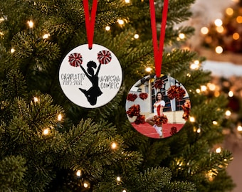 May include: Two round ceramic ornaments with red ribbon hangers. One features a black silhouette of a cheerleader with pom-poms and the text "Charlotte 2025-2026 Warsaw Comets". The other displays a photographic image of a cheerleader in uniform.