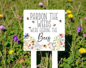 May include: A white rectangular garden sign with two stakes featuring the black text "PARDON THE WEEDS WE'RE FEEDING THE Bees" and a colorful illustration of various wildflowers and small bees scattered across the surface of the sign.