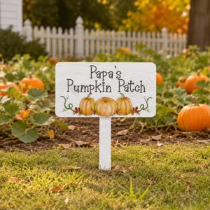May include: A rectangular white garden sign with a wood grain texture featuring the black text Papa's Pumpkin Patch and an illustration of three orange pumpkins with green vines and small red leaves, mounted on a matching white stake.