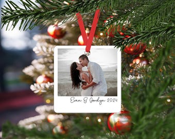 May include: A square white ornament featuring a photograph of a couple kissing on a beach. The ornament has a red ribbon for hanging and includes the printed text Evan & Jordyn 2024 at the bottom, styled like a classic instant film photograph.
