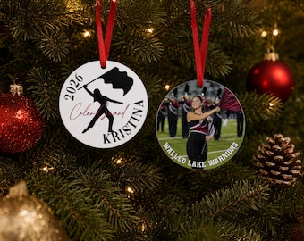 May include: Two round white ornaments with red ribbon hangers. The left ornament features a black silhouette of a colour guard member, the text 2026, Colour Guard, and KRISTINA. The right ornament displays a photo of a colour guard member and WALLED LAKE WARRIORS.