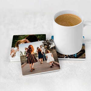 May include: A set of three square coasters featuring printed photographs of a family walking on a wooden bridge, accompanied by a white ceramic mug filled with coffee. The coasters have a smooth finish and display outdoor scenes with people.