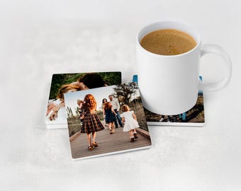 May include: A set of three square coasters featuring printed photographs of a family walking on a wooden bridge, accompanied by a white ceramic mug filled with coffee. The coasters have a smooth finish and display outdoor scenes with people.