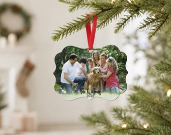 May include: A custom-shaped holiday ornament featuring a printed photograph of a family with a golden retriever dog. The ornament has a scalloped edge, a red ribbon for hanging, and the black text The Wilsons 2026 printed at the bottom.
