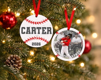 May include: Two round, white, flat ornaments with red ribbon hangers. One features red baseball stitching, the name CARTER, and the year 2026 in gray. The other displays a photograph of a baseball player and the text COMMERCE HAWKS in gray.