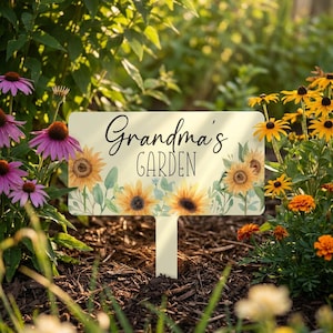 May include: Cream-colored garden sign with the words "Grandma's GARDEN" in black script. The sign features watercolor sunflowers and greenery. The sign is staked in a garden bed surrounded by flowers.