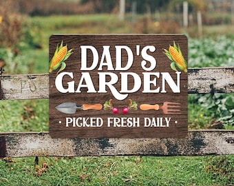 May include: A rectangular sign with a dark brown wood grain background featuring the white text DAD'S GARDEN and PICKED FRESH DAILY. The design includes illustrations of two yellow corn cobs, a metal trowel, two red radishes, and a brown garden fork.