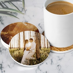 May include: Two round coasters featuring a printed photograph of a couple embracing in a forest setting with tall trees and tall grass. One coaster is placed under a white ceramic mug filled with coffee, while the other is partially visible.