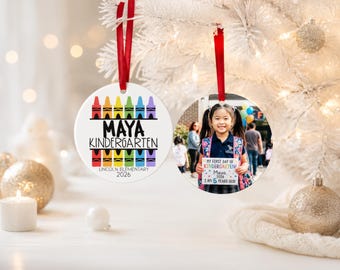 May include: Two round white ceramic ornaments with red ribbon hangers. One features colorful crayon graphics, the name Maya, Kindergarten, Lincoln Elementary, and 2026. The other displays a photograph of a child holding a sign reading My First Day of Kindergarten, Maya, 2026, I Am 5 Years Old.