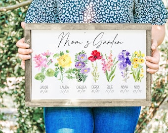 May include: A framed wooden sign with the words "Mom's Garden" in elegant script. The sign features watercolor illustrations of various flowers, each labeled with a name and date. The flowers are in shades of pink, yellow, purple, and red.