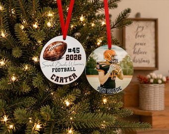 May include: Two round white ornaments hanging by red ribbons. One features a brown football illustration, the text #45 2026, Sarah Banks Warriors FOOTBALL, and CARTER. The other shows a football player in a black and orange uniform with 9TH GRADE.