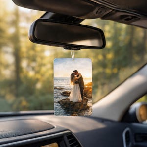 May include: A rectangular car air freshener hanging from a rearview mirror, featuring a printed photograph of two people embracing on a rock by the ocean at sunset, with warm golden light reflecting off the water and the sky in the background.