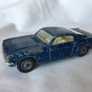 May include: A blue toy car model of a Ford Mustang with a white stripe along the top. The car has a weathered, worn look, with some paint missing.