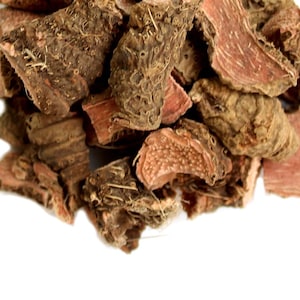 Może przedstawiać: Stos suszonych, grubo pociętych kawałków czerwonobrązowego korzenia, prawdopodobnie ziołowego środka leczniczego lub przyprawy. Kawałki mają szorstką, teksturowaną powierzchnię zewnętrzną i czerwoną wewnętrzną, na białym tle.