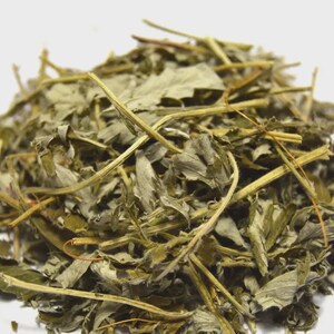 May include: A close-up of dried green leaves and stems, possibly herbs or tea. The leaves are various sizes and shapes, with a mix of light and dark green hues. The stems are thin and straw-colored, creating a textured appearance against a white background.