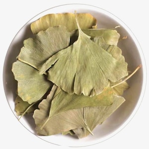 May include: A white bowl filled with dried ginkgo biloba leaves. The leaves are a muted green color with a fan-like shape and prominent veining. The bowl is round and white, contrasting with the leaves.