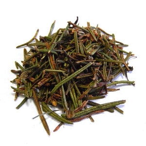 May include: A close-up of a pile of dried herbal tea leaves, predominantly green and brown in colour. The leaves have a needle-like form and are scattered against a white backdrop. The tea leaves appear to be a natural product.