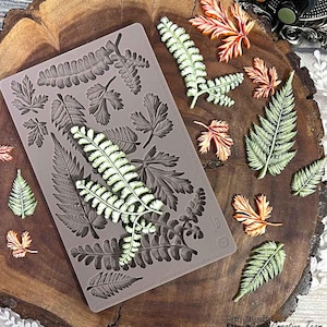May include: A gray silicone mold with various leaf designs, alongside several painted leaf embellishments in shades of green, orange, and white. The mold features detailed botanical patterns, perfect for crafting projects. The leaves are arranged on a wooden surface.