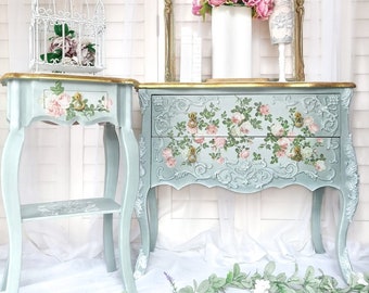 May include: Two vintage-style side tables with a light blue finish and floral designs. The larger table has two drawers and the smaller table has a shelf. Both tables have gold accents and are decorated with pink roses.