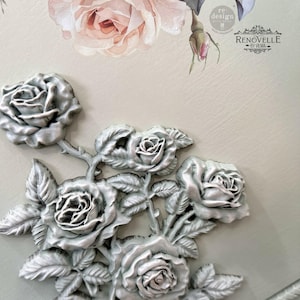 May include: A detailed close-up of a decorative panel featuring a 3D floral design. The design showcases a cluster of silver-colored roses with intricate detailing, set against a pale green background. The panel also includes a floral decal and the text "RENOVELLE BY VILMA".