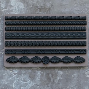 May include: A collection of black decorative molding designs, including rope, beaded, and floral patterns, arranged on a rectangular, light brown surface. The designs are set against a textured, gray background.
