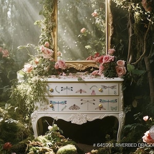 May include: A white dresser with two drawers decorated with dragonfly and floral designs. The dresser is in a forest setting with a gold mirror and pink roses.
