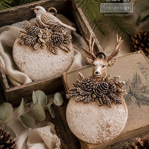 May include: Two round, textured, off-white ornaments featuring raised relief designs. One ornament displays a bird perched atop pinecones and foliage, while the other features a deer head surrounded by pinecones and evergreen branches.