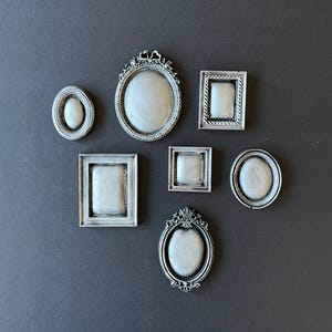 May include: A collection of seven small, silver-colored picture frames in various shapes: oval, rectangular, and square. The frames have ornate detailing and are arranged against a dark gray background. The frames are empty.