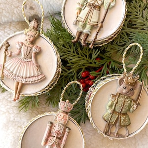 May include: Four round Christmas ornaments featuring a Nutcracker, a ballerina, a mouse, and a Nutcracker. The ornaments are white with gold trim and feature detailed illustrations of the characters. The ornaments are hanging from a Christmas tree.