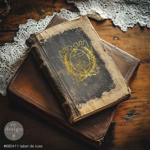 May include: Two antique books with distressed covers rest on a wooden surface. The top book has a dark cover with a gold emblem and text. The bottom book is brown. The image includes the text "#680411 label de luxe".