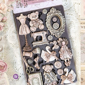 May include: A dark gray silicone craft mold labeled buttons & keepsakes, featuring recessed shapes of a dress form, teddy bears, an ornate mirror, a sewing machine, a swan, a doll, scissors, a spool of thread, buttons, and a small book.