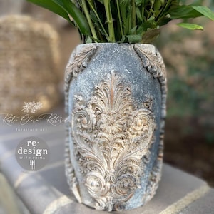 May include: A gray ceramic vase with an ornate, raised design in a light brown color. The vase is decorated with a distressed finish and has a slightly textured surface.