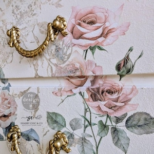 May include: A white dresser with two drawers featuring a floral design with pink roses and green leaves. The dresser has gold metal drawer pulls with a rope design. The dresser is labeled "re-design with grit" and "shabby chic & co. restored furniture".