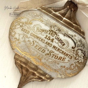May include: Ornament with a vintage design. The ornament is off-white with gold lettering that reads "Flower and Garden Seed Store." The ornament has a decorative shape and a ribbon for hanging.