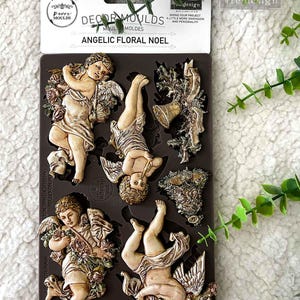 May include: A dark brown silicone mold featuring detailed angel and floral designs. The mold includes multiple cherub figures with wings, flowers, and a bell. The packaging reads "ANGELIC FLORAL NOEL" and "DECOR MOULDS".