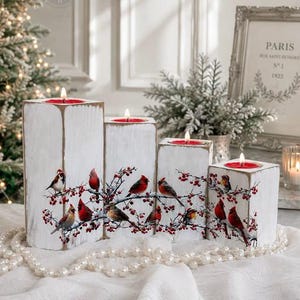 May include: A set of four rectangular white wooden candle holders with distressed edges, each featuring a printed design of red cardinals perched on snowy branches with red berries. Each holder contains a single lit red pillar candle.