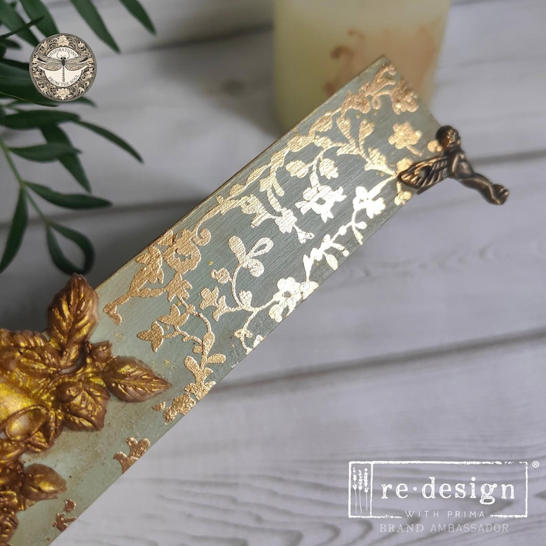 May include: A rectangular wooden bookmark featuring a sage green base with a metallic gold floral pattern. The bookmark is adorned with a three-dimensional gold leaf embellishment at the bottom and a small bronze-toned metal angel figure at the top.