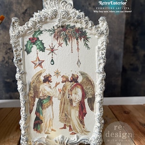 May include: Ornate, white framed artwork featuring a Christmas scene with angels and stars. The artwork has a distressed finish and includes the text "Retro Interior" and "re design with prima". The frame is approximately 12 inches tall.