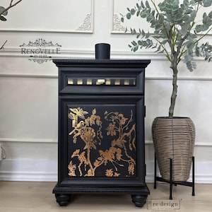 May include: Black cabinet with gold floral design and a small bird. The cabinet has a rectangular shape with a decorative top and sits on four rounded feet. A black candle sits on top. The cabinet is next to a potted plant.