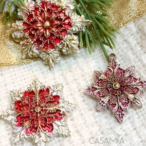 May include: Three ornate snowflake ornaments with a red, gold, and white color scheme. The ornaments are made of metal and have a detailed design. The ornaments are on a white background.