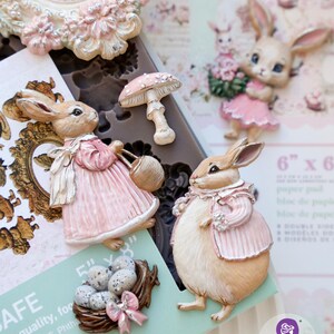 May include: A collection of decorative resin bunny figurines, a mushroom, and a nest with eggs. The bunnies wear pink dresses and jackets. The frame and the nest are decorated with pink bows. The background includes a floral pattern.