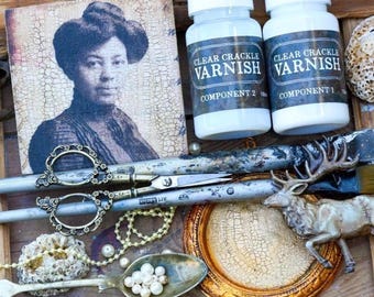 May include: Two white plastic bottles labeled Clear Crackle Varnish Component 1 and 2, a square print of a person in a hat, ornate metal scissors, a metal spoon with white beads, a gold circular frame, and a metallic deer figurine on wood.