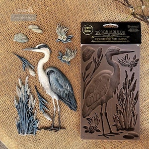 May include: A collection of decorative elements featuring a heron, including a painted heron figure, small stones, and floral accents. A package of "Feathered Stillness" decor molds is also present, showcasing a heron design.