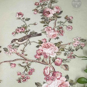 May include: A decorative transfer featuring a brown bird perched on a branch with pink and white flowers and green leaves against a light sage green background. The text re: design with prima is visible in the upper right corner of the design.
