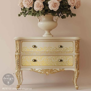 May include: A cream-colored two-drawer nightstand with gold accents and floral detailing. The drawers feature a yellow floral pattern and gold knobs. A vase of pink roses sits atop the nightstand. The text on the bottom reads "#676155 vineyard elegance".