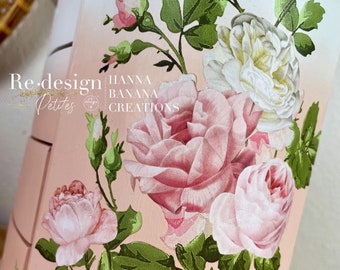 Puede incluir: Una cómoda pintada de rosa con tres cajones presenta un diseño floral con rosas rosas y blancas y hojas verdes. El texto "Re-design Petites" y "HANNA BANANA CREATIONS" está impreso en la cómoda.