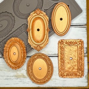 May include: A collection of six ornate, gold-colored decorative elements. The pieces include oval and rectangular shapes with detailed, vintage-style frames. Each piece has a central hole, suggesting they are designed for attachment.