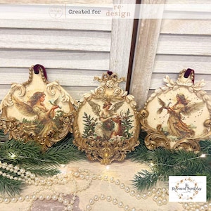 May include: Three ornate, cream-colored Christmas ornaments with gold filigree frames. Each features a vintage-style angel illustration. The ornaments have burgundy velvet ribbon hangers and are arranged on a bed of greenery and pearls.