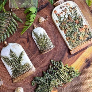 May include: Decorative resin cloches and botanical accents. The image features three cloches with fern and floral designs, along with a cluster of green fern leaves and small snail figurines. The cloches have a distressed, antique finish, and the overall theme is nature-inspired.